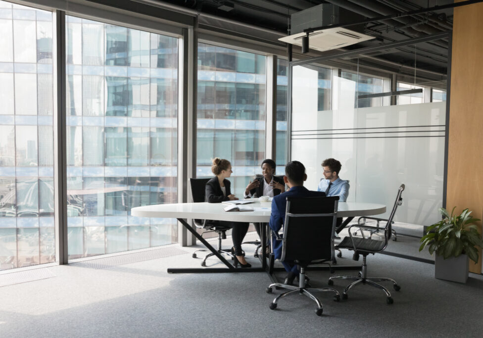 Diverse millennial business team talking in meeting room, negotiating on project at table at glass wall panoramic window, discussing deal in open space, modern office interior. Wide shot